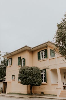 Beautiful two-story house with green shutters and lush trees in a serene setting.