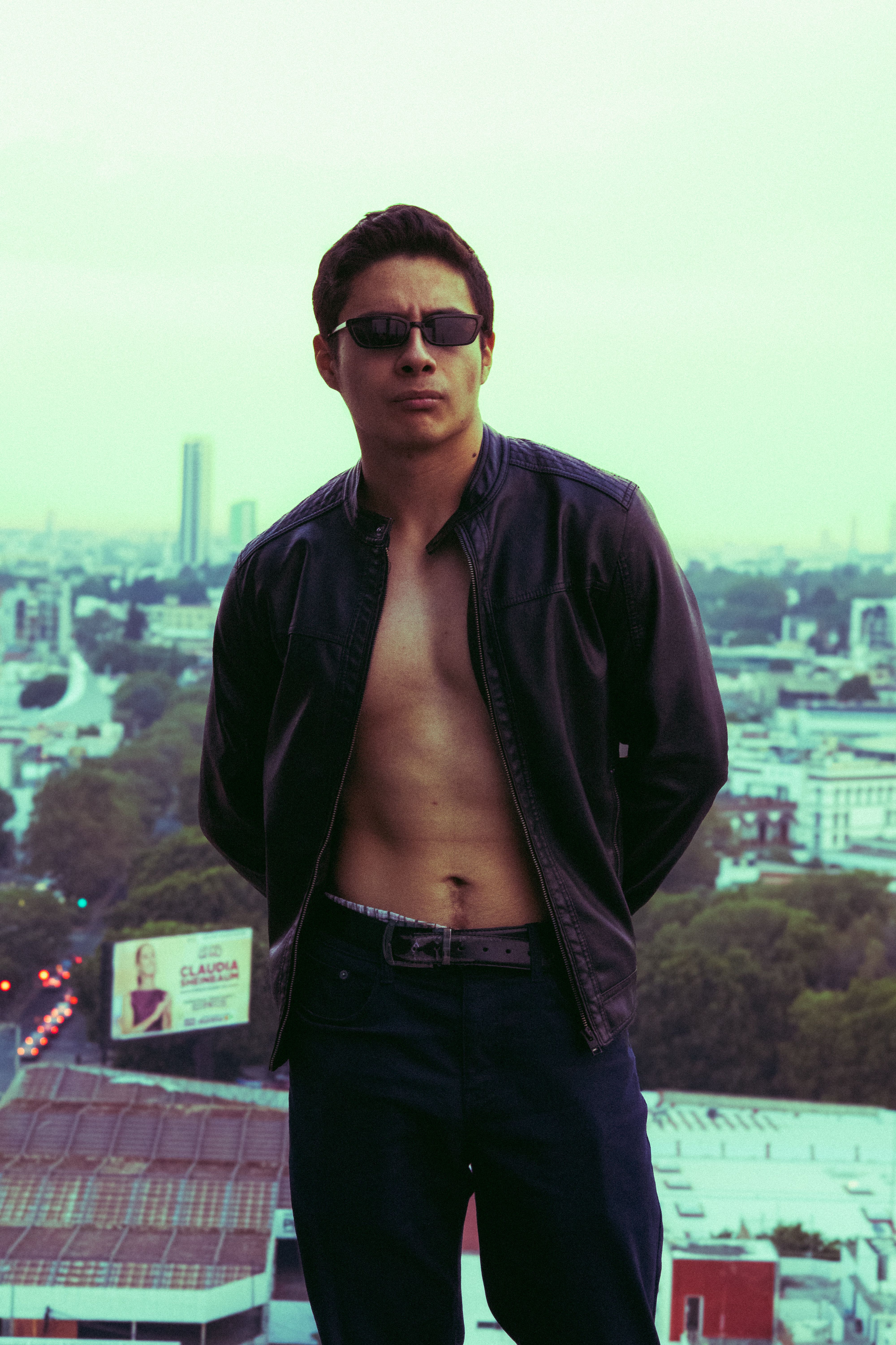 Portrait of a shirtless young man in sunglasses and black jacket, standing against an urban backdrop.