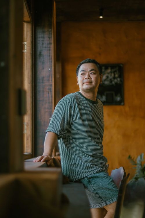 Free Man in a striped shirt leaning on a sill, gazing out a window in a cozy room. Stock Photo