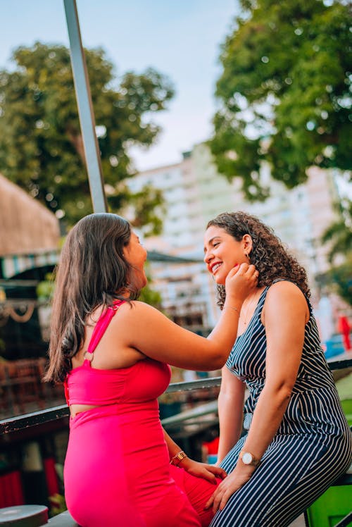 Two women sitting on a bench and one is putting on the other's face