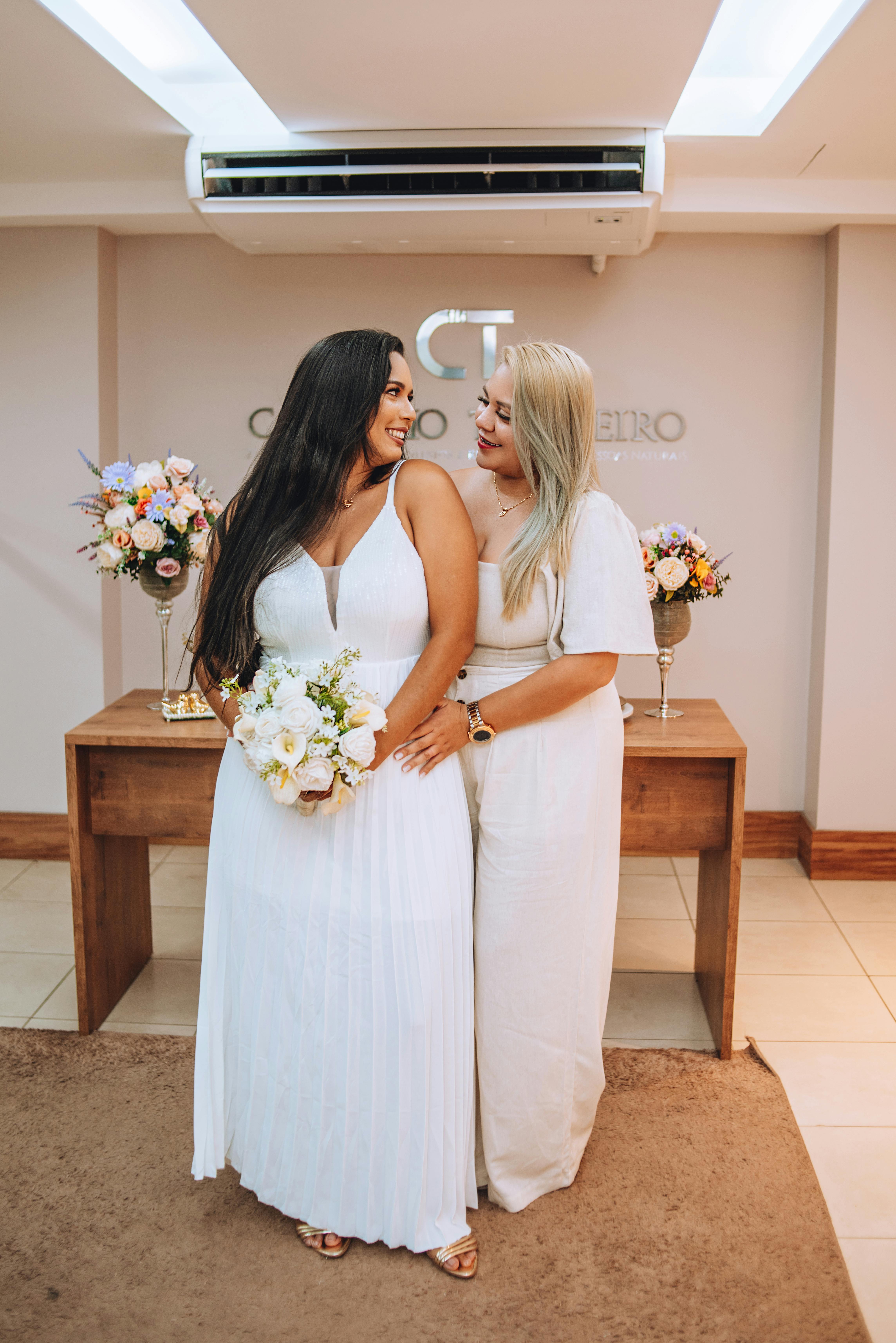 Two women in white wedding dresses smiling and embracing indoors with floral decor.