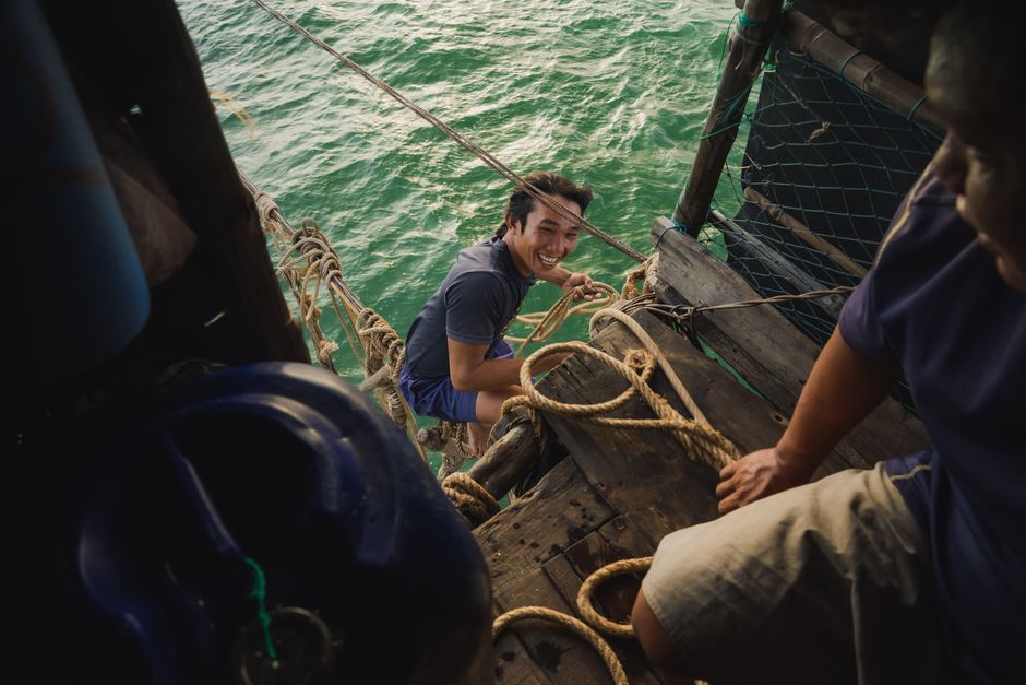 Man climbing ropes on a wooden structure by the sea, smiling at the camera, adventurous outdoor activity.