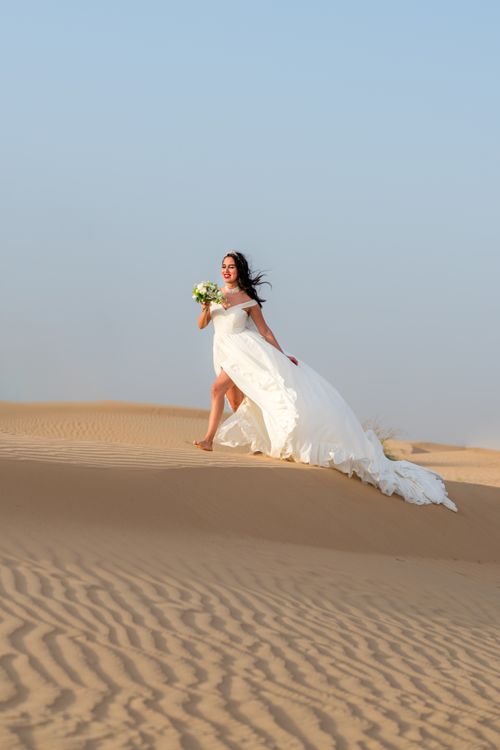 Free A bride in a white wedding dress walks through the Dubai desert holding a bouquet. Stock Photo