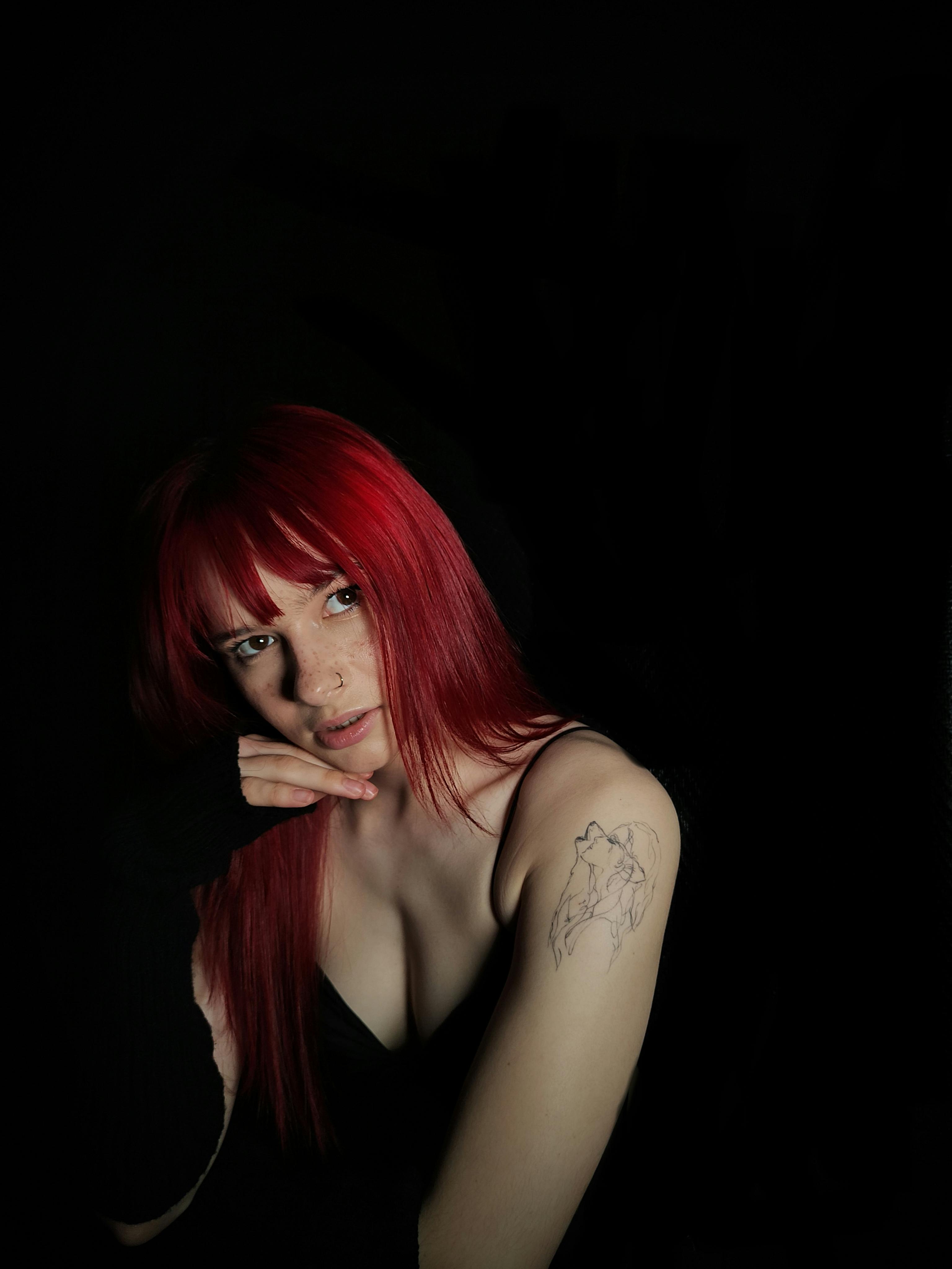 Artistic studio portrait of a woman with vibrant red hair and a tattoo, captured against a dark background. Perfect for fashion or editorial use.