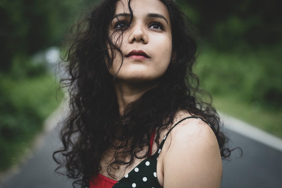 A contemplative woman with curly hair gazes upwards on a road surrounded by greenery.