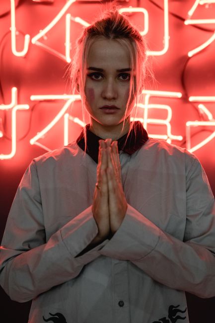 Stylish woman standing with clasped hands in front of a red neon sign, serious expression.