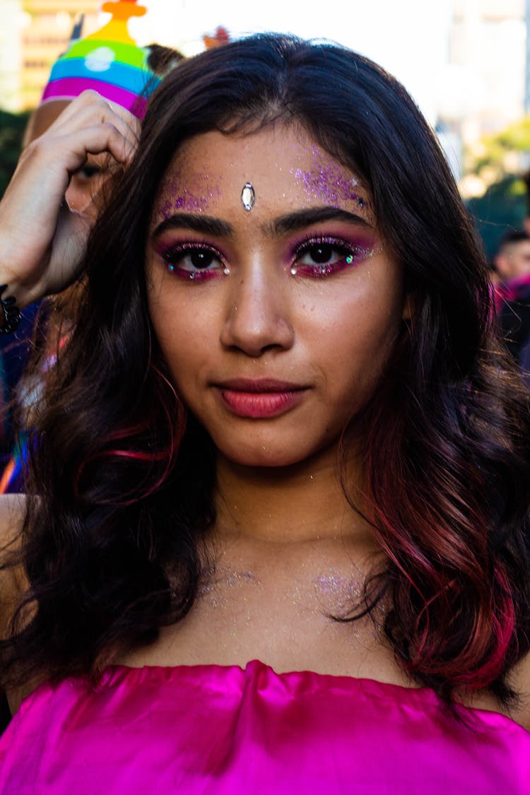 Portrait Photo Of Woman On Pink Off-shoulder Top And Pink Glitter On Her Face Posing With Crowd Of People In The Background