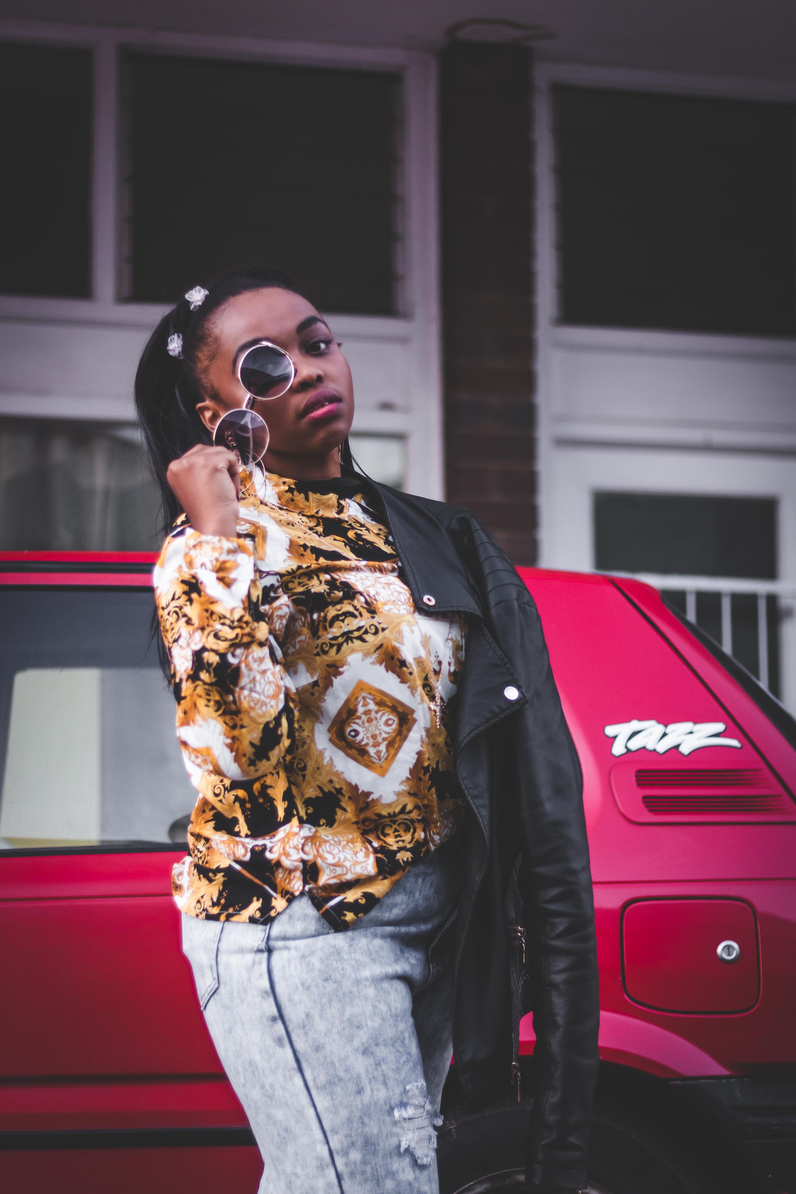 Uma mulher fashion usando óculos de sol e roupas casuais posando confiantemente ao lado de um carro vermelho ao ar livre.
