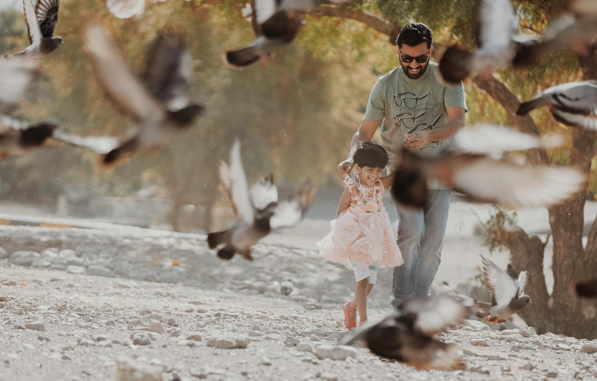 Father and daughter joyfully play among pigeons in a sunny outdoor setting, capturing family happiness.