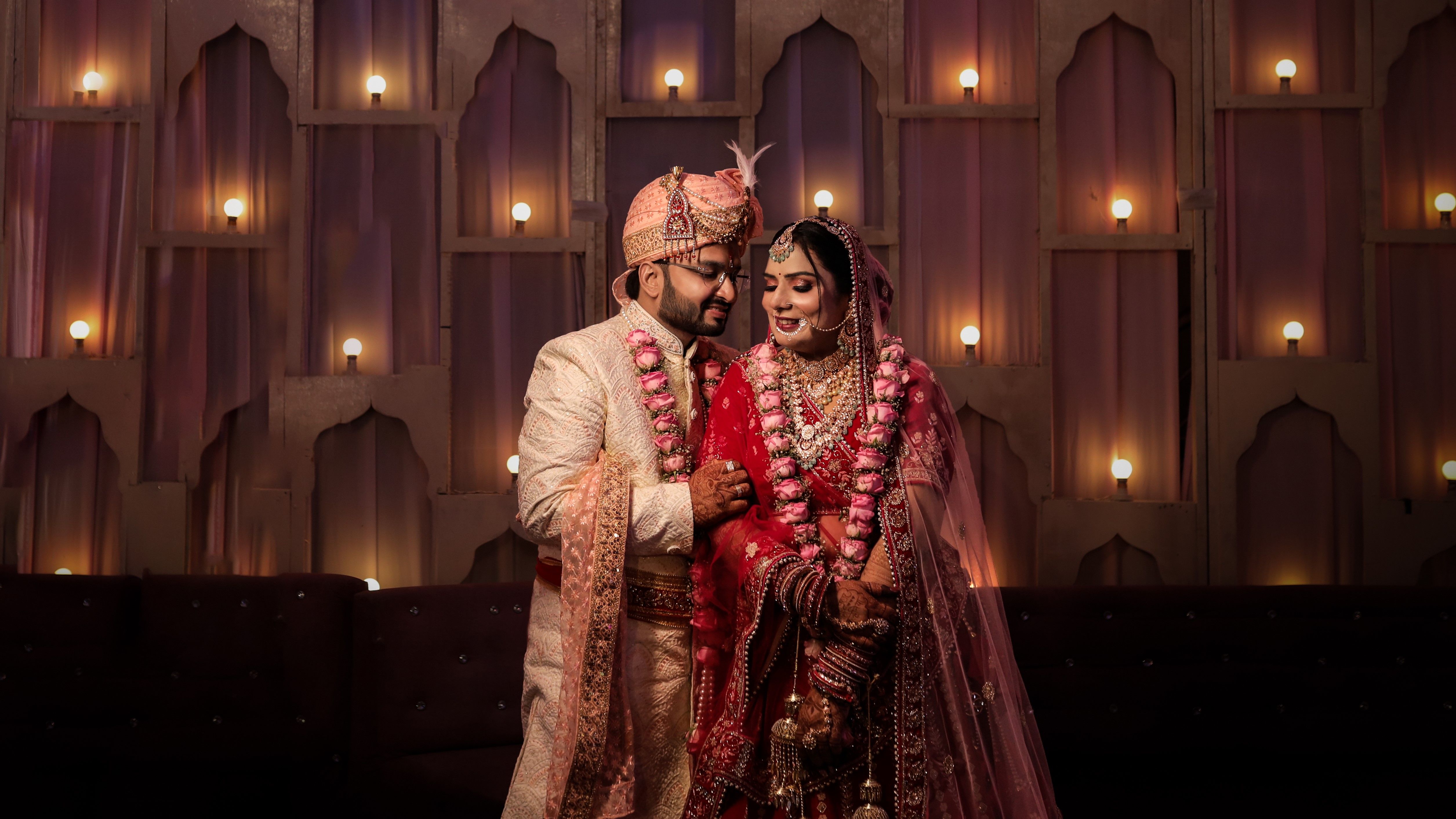 Indian newlywed couple in traditional clothing, smiling and holding hands indoors.