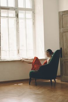 A woman in a red dress sits pensively by a bright window in a stylish room.