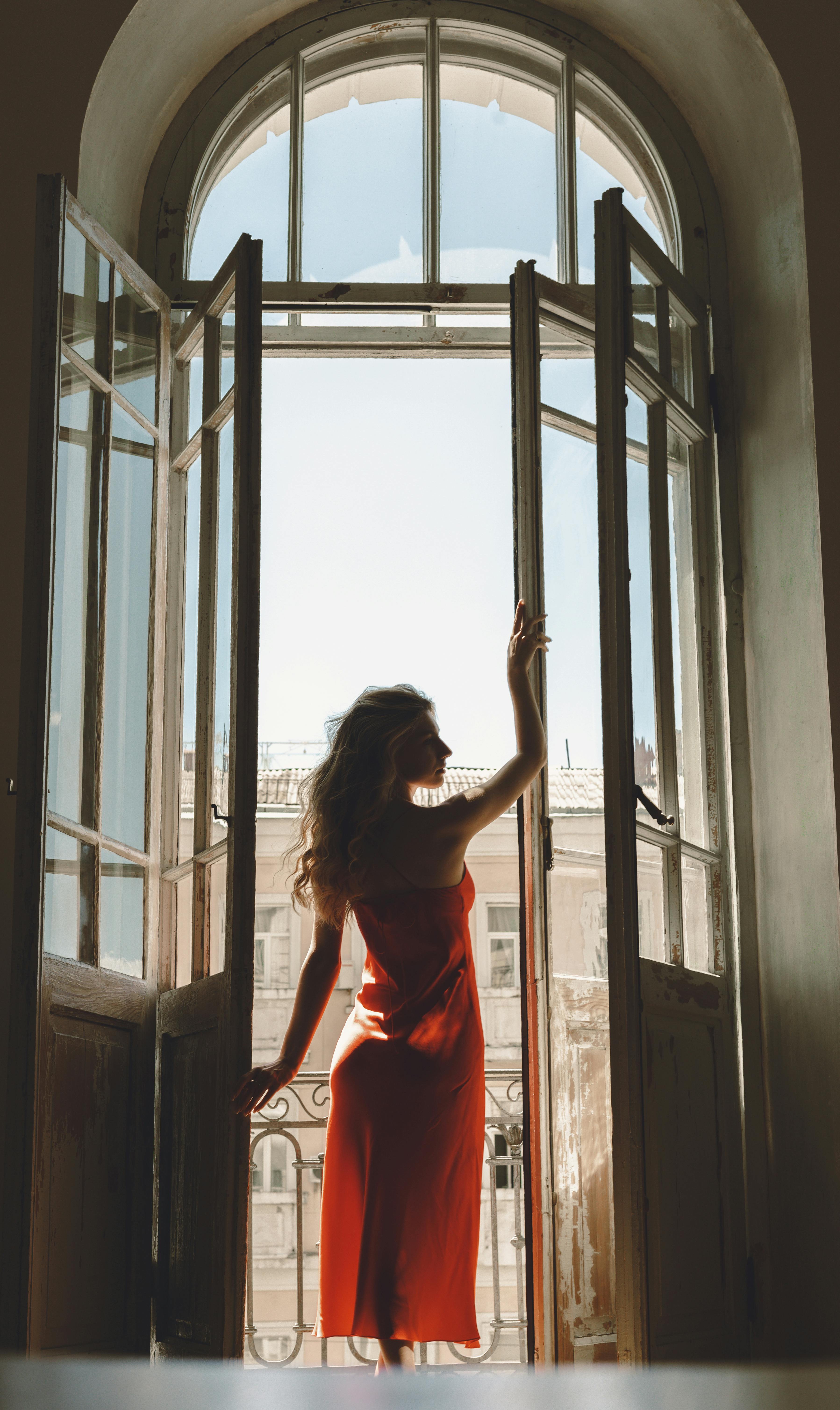 Silhouette of a woman in a red dress standing by an open balcony door at day.