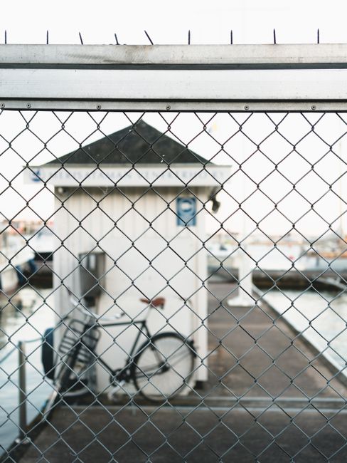 Chain link fence framing a dock scene with a bicycle near a small shelter by the water.