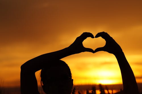 Free Silhouette Woman Hand Holding Heart Shape Against Orange Sky Stock Photo