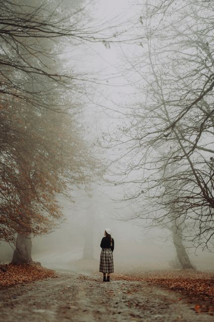 A woman walks on a foggy dirt road surrounded by autumn trees in a mysterious forest.