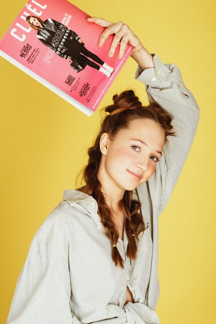 Portrait of a smiling woman with braided hair holding a magazine against a yellow background.