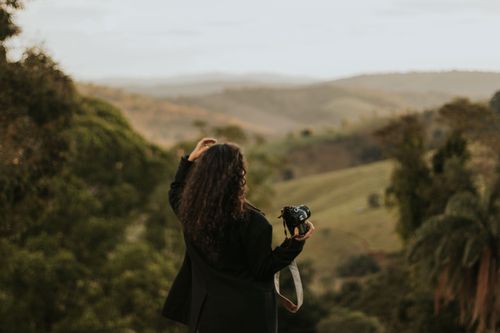 Free Back view of a woman photographer with long curly hair, capturing a scenic landscape of rolling hills. Stock Photo