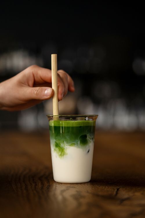 Free A person's hand stirring a refreshing iced matcha latte with a bamboo straw on a wooden table, captured in a cozy setting. Stock Photo