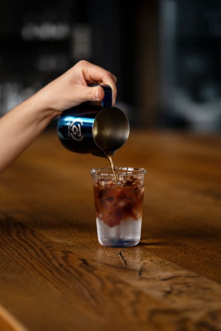 Hand pouring coffee from a metallic pitcher into a glass on a wooden surface, captured with a selective focus.