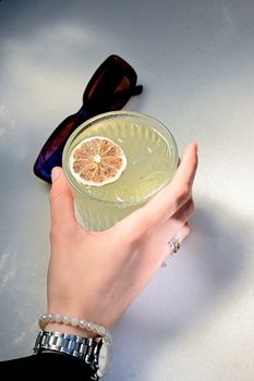 Hand holding a cold lemonade garnished with dried lemon slice next to sunglasses on a sunny day.