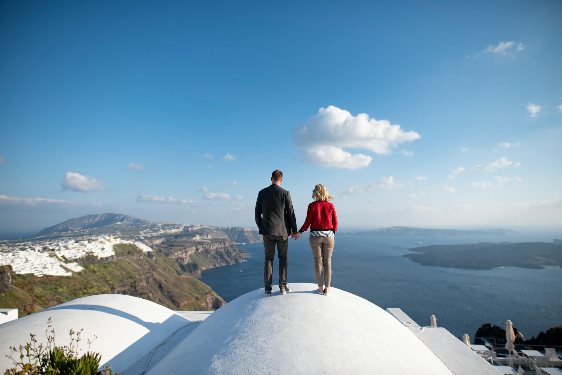 A couple stands hand-in-hand on a rooftop with a stunning view of Santorini and the Aegean Sea.