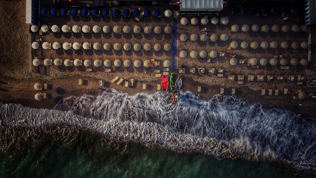 Aerial view of a beach with aligned umbrellas, waves gently hitting the shore, and sunlight casting shadows.