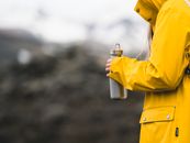 一个穿着黄色雨衣的人，手持金属水瓶，映衬在模糊的冰岛风景中