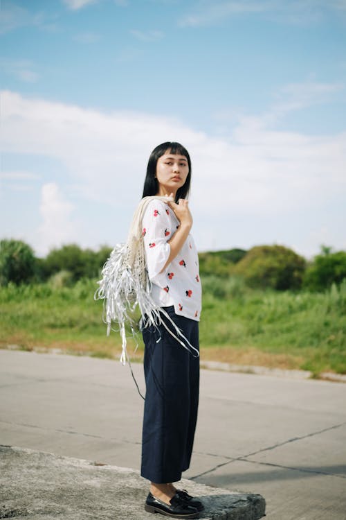 Free Side View Photo of Woman in White and Red Blouse and Blue Pants Standing on Concrete Surface Posing While Carrying Bag Stock Photo