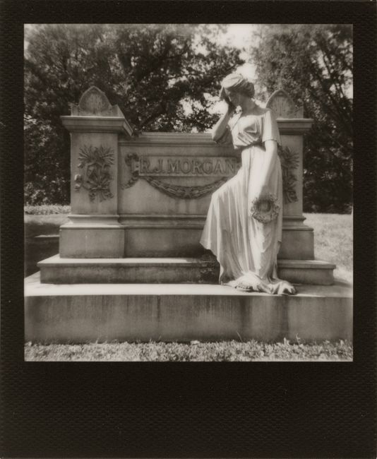 Black and white Polaroid of a statue beside a gravestone in a serene outdoor setting.