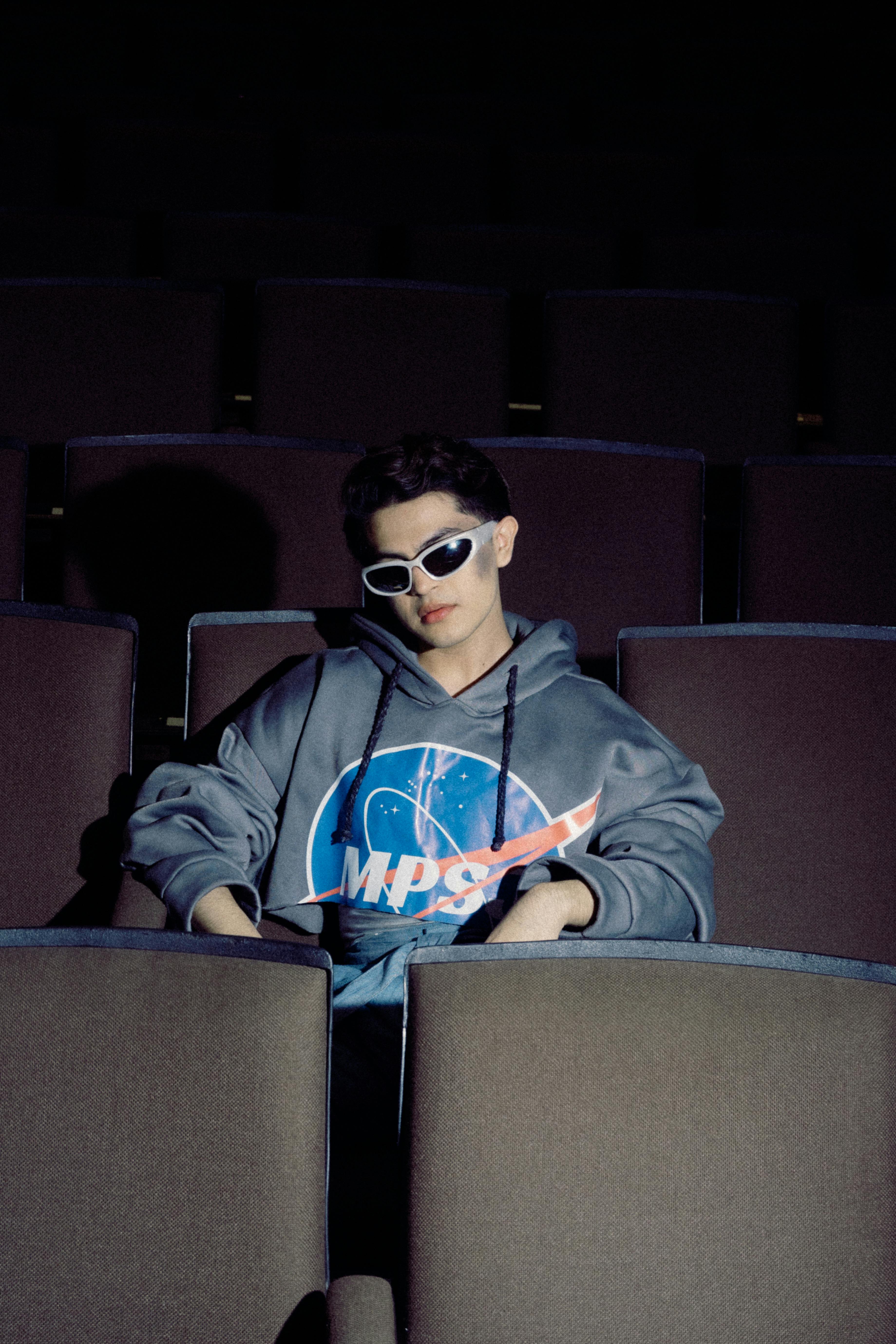 A stylish individual in sunglasses sits casually in a dimly lit movie theater.