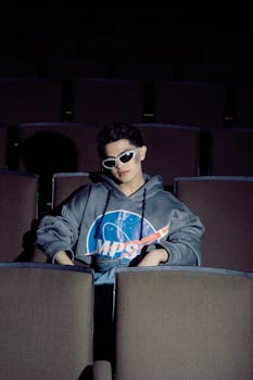 A stylish individual in sunglasses sits casually in a dimly lit movie theater.