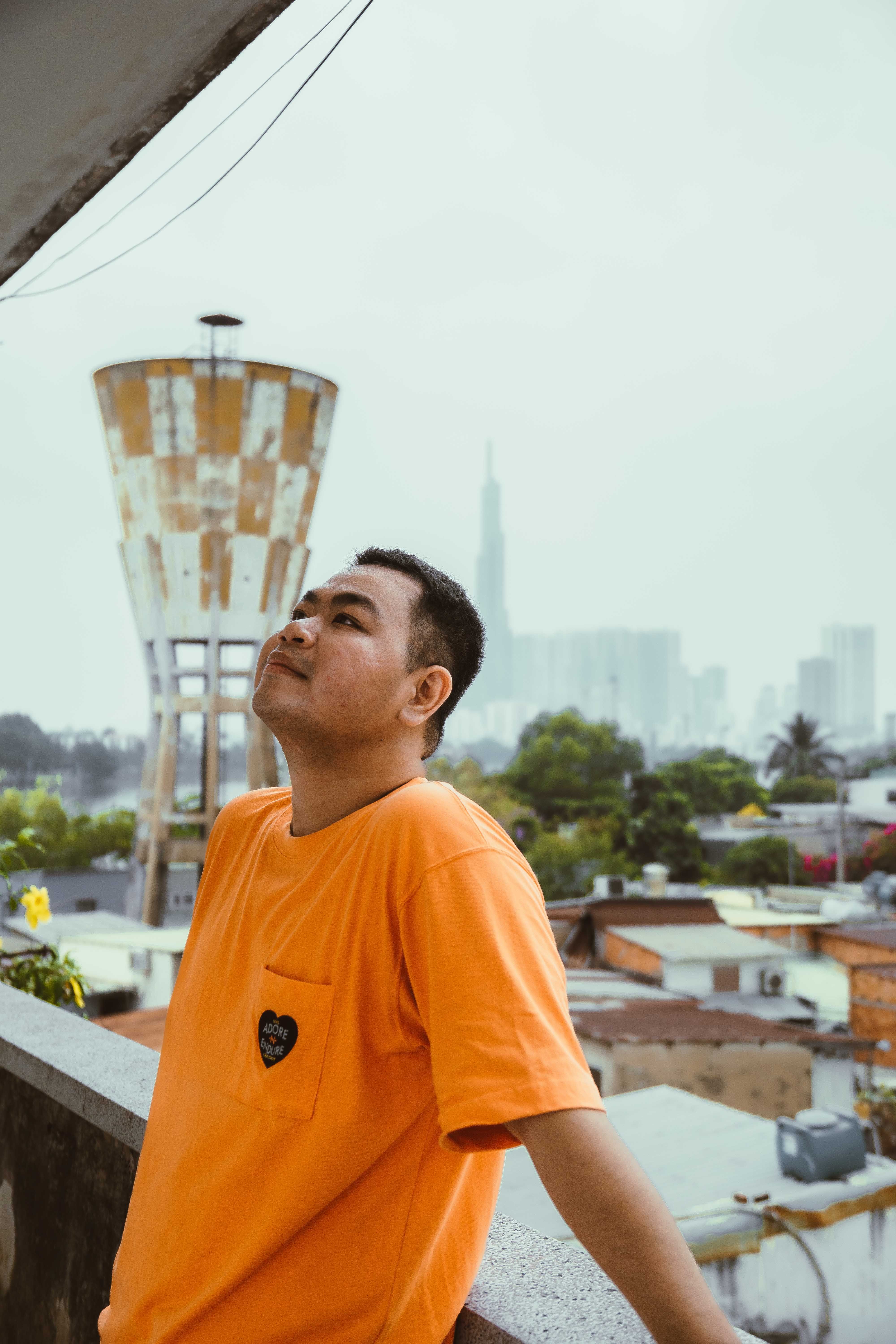 Man in orange shirt standing on balcony with urban skyline and water tower in background.