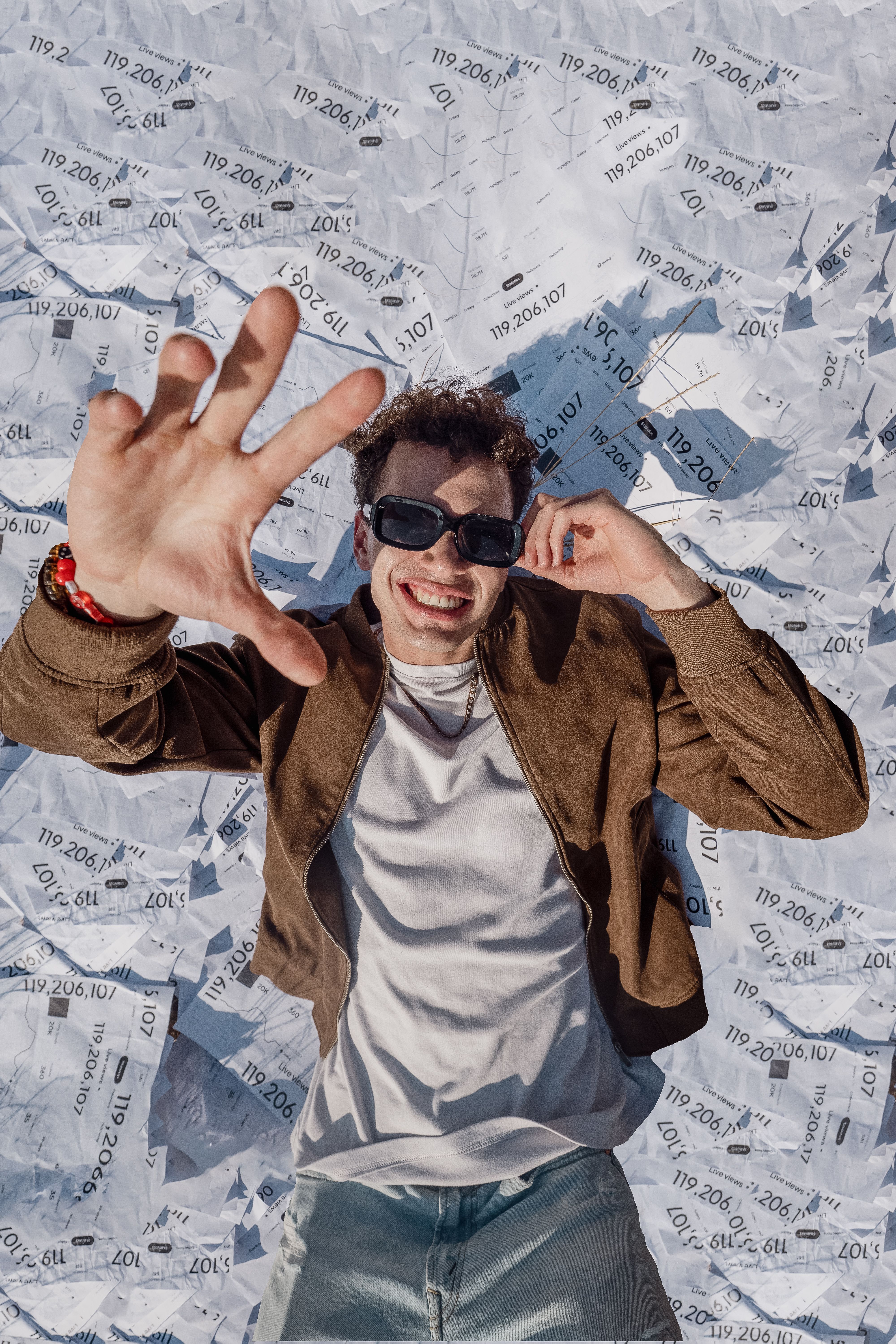 Smiling young man wearing sunglasses, lying on a pile of papers, outdoors.