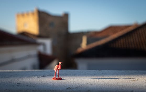 A small toy flamingo atop a rooftop against a blurred urban backdrop at sunset in Sines, Portugal.