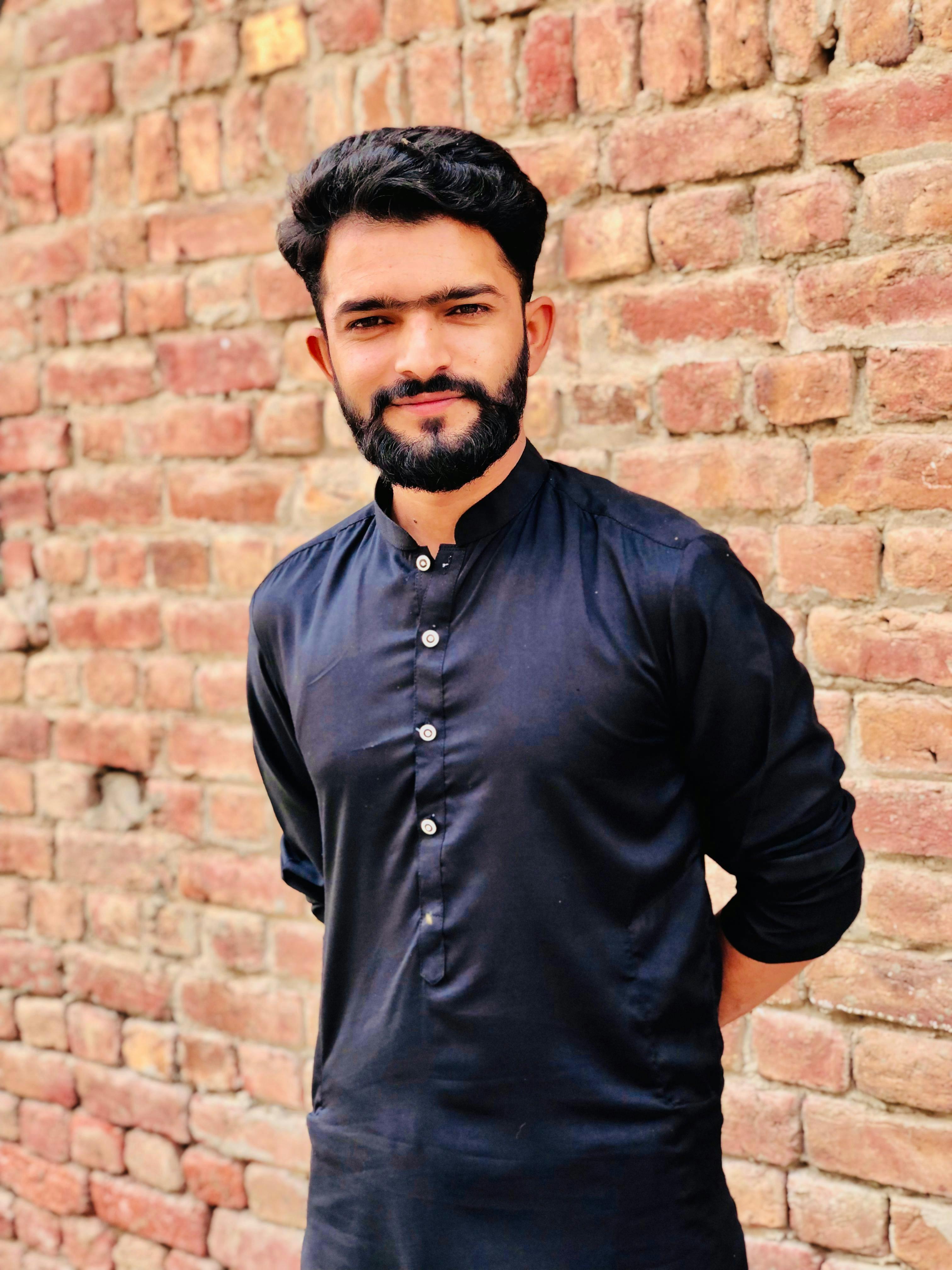 A confident man in a black kurta stands against a rustic brick wall, exuding style.