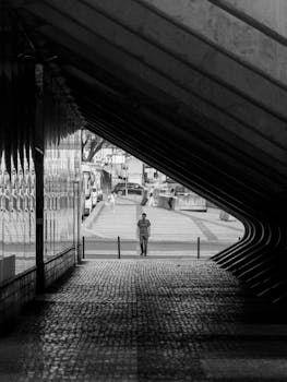 A solitary figure stands in a modern urban passageway, captured in striking black and white tones.
