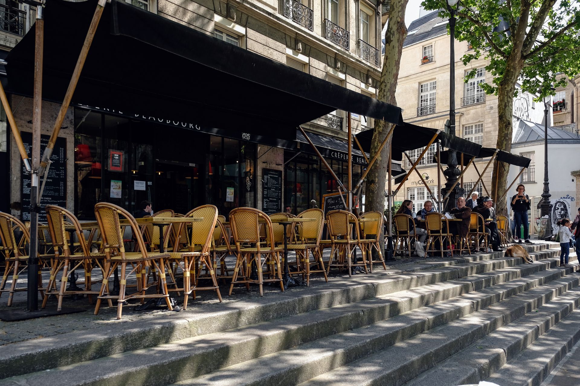 Outdoor Paris café with wicker chairs and people enjoying a sunny day on stone steps.