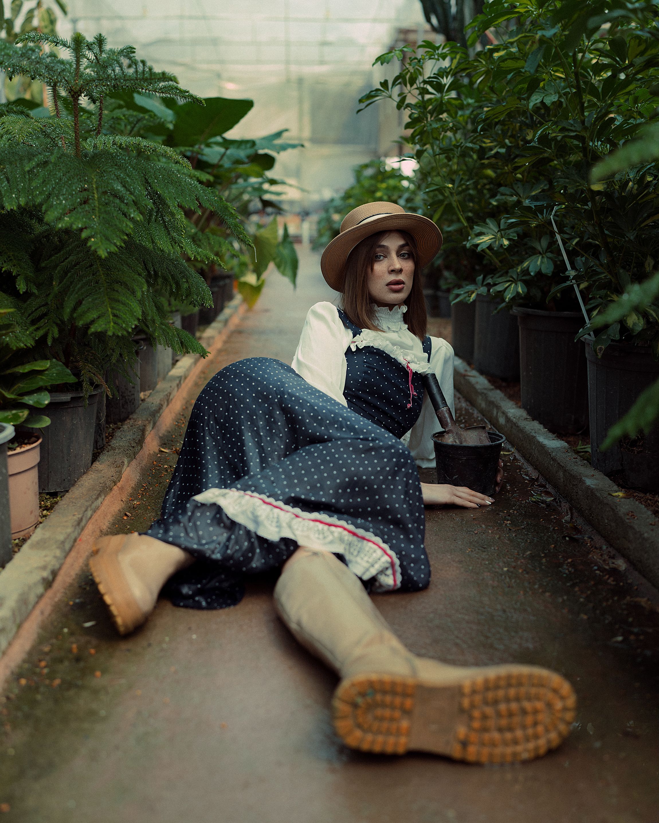 Woman in a vintage dress and hat poses in a verdant greenhouse setting.