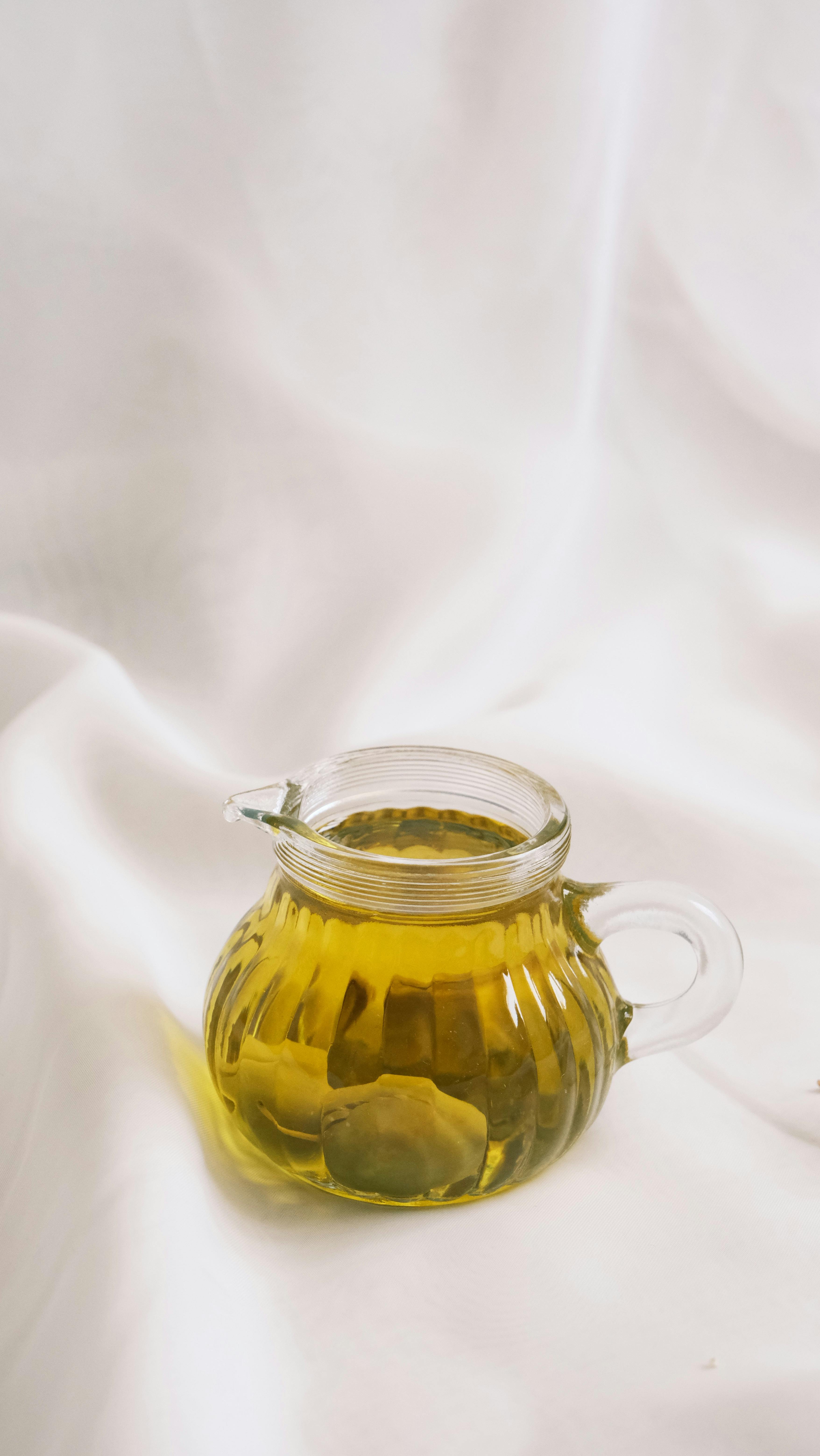 A clear glass pitcher filled with olive oil, set against a soft white fabric backdrop.