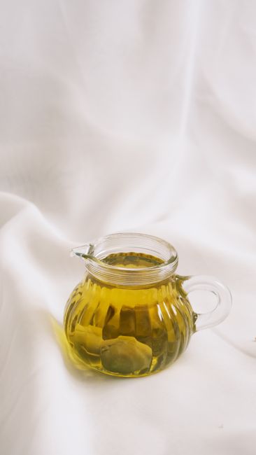 A clear glass pitcher filled with olive oil, set against a soft white fabric backdrop.