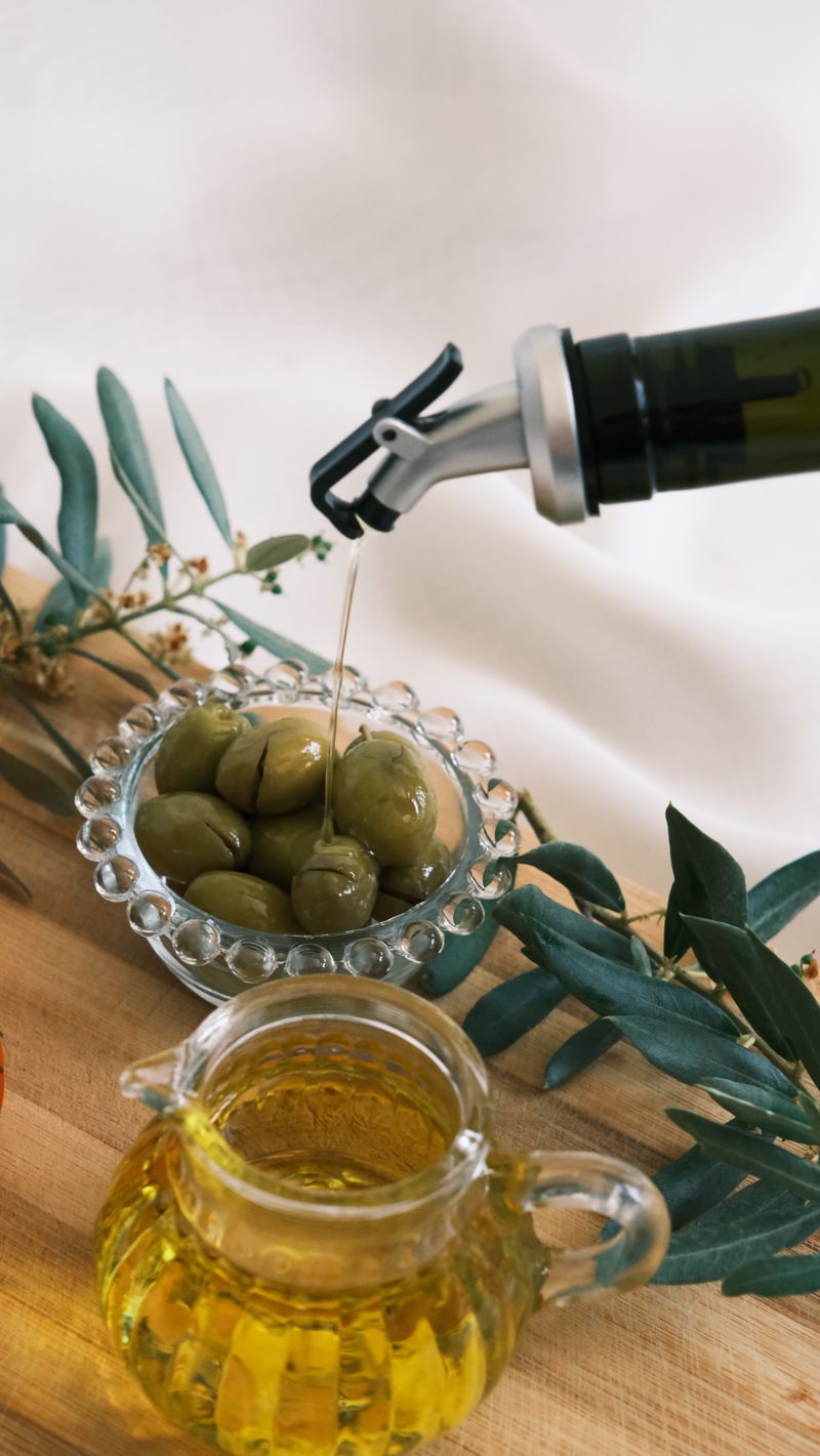 Olive oil pouring over a bowl of green olives next to olive branches