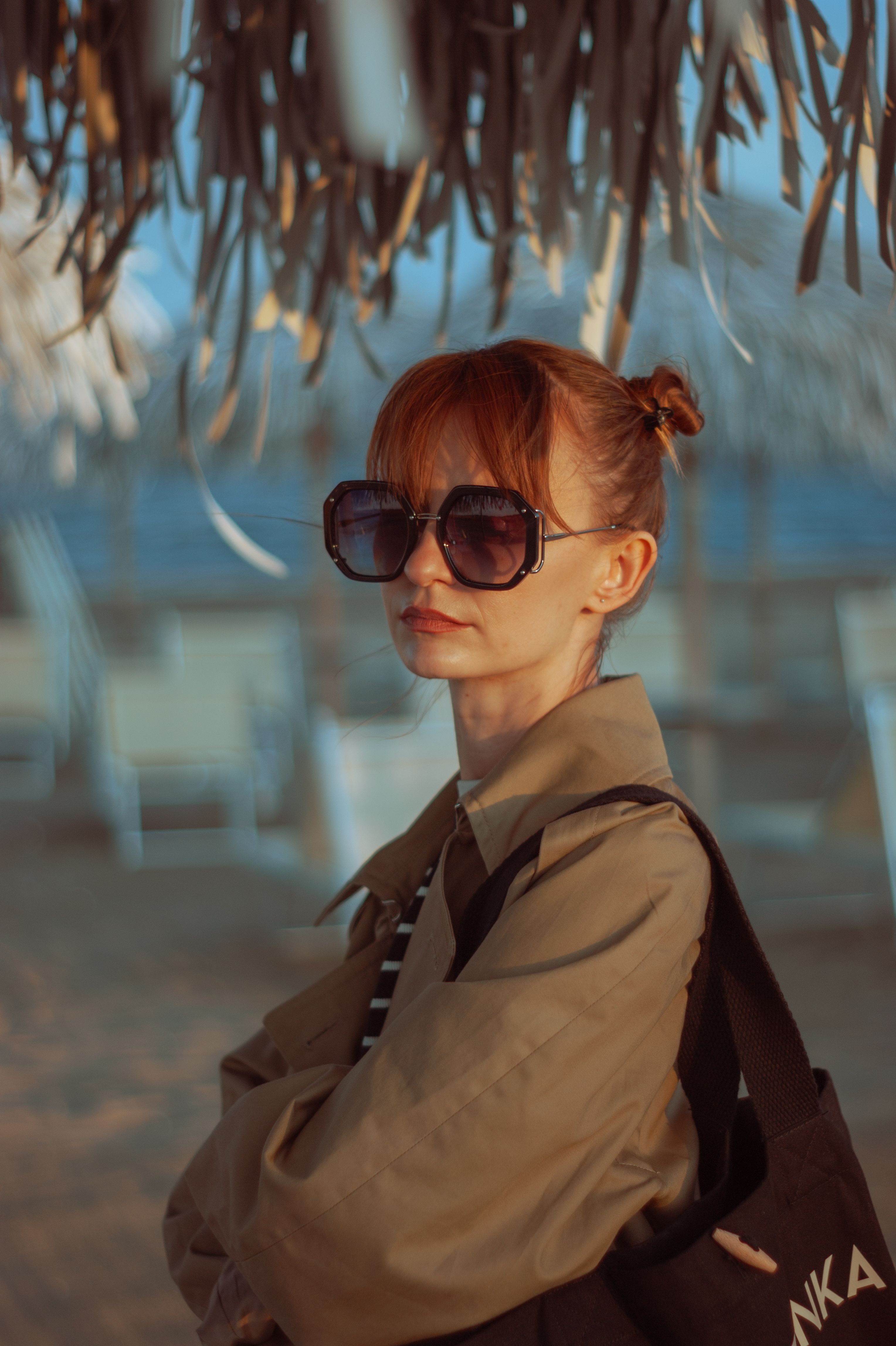 Chic woman with sunglasses and brown hair posing under a palm hut at the beach.