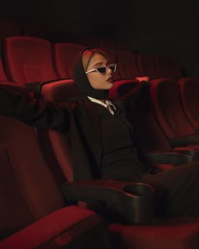Fashionable woman in black attire wearing sunglasses, seated elegantly in a dimly lit cinema.