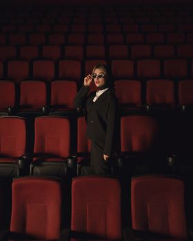 Elegant woman poses in a black suit and sunglasses in an empty cinema with red seats.