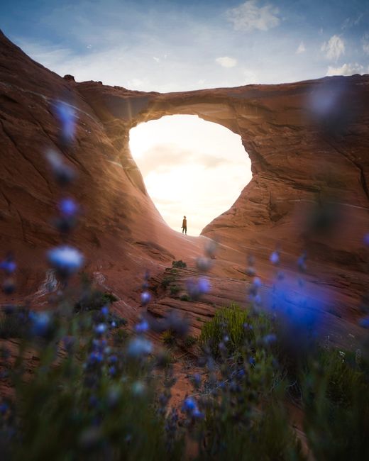 A lone figure stands silhouetted under a beautiful natural canyon arch at sunset.