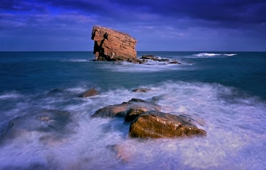 A striking view of rocks and waves crashing under a dramatic blue sky at sea.