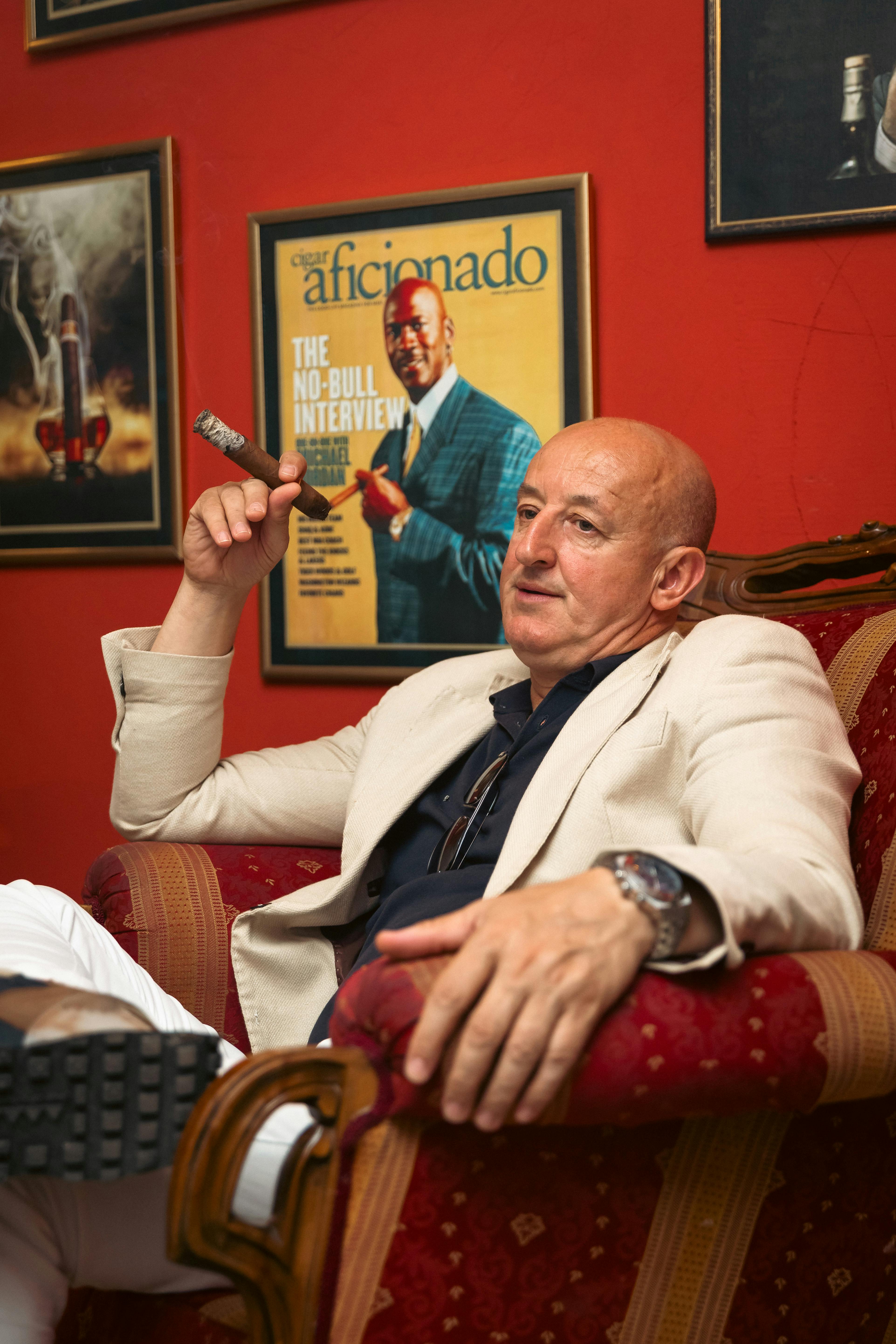 A bald man in a suit sits in an armchair smoking a cigar, exuding elegance and relaxation.