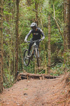 An adult mountain biker jumps over a dirt trail in a dense forest, showcasing dynamic action and adventure.