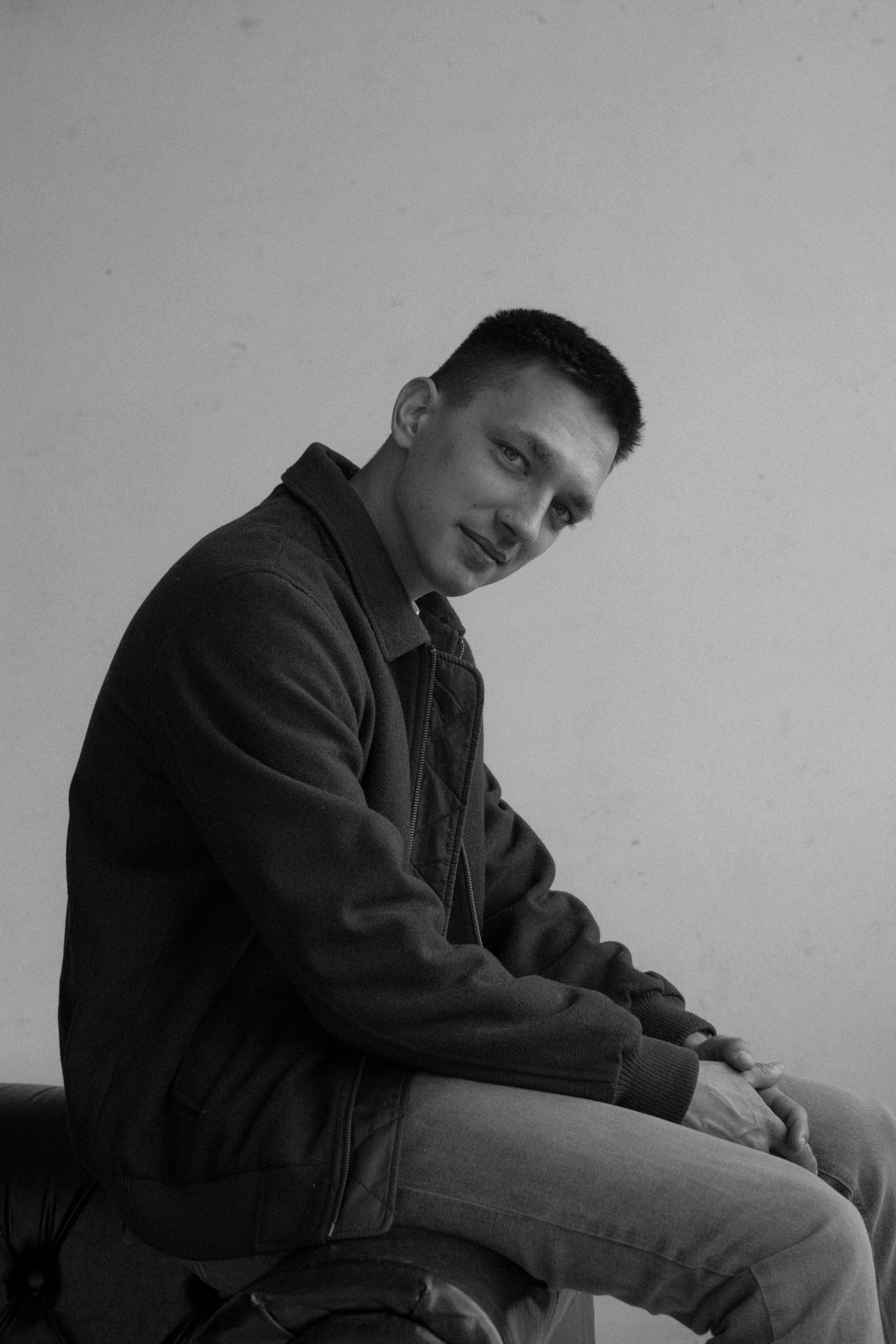 A stylish black and white portrait of a man sitting and smiling, wearing casual attire with a jacket.