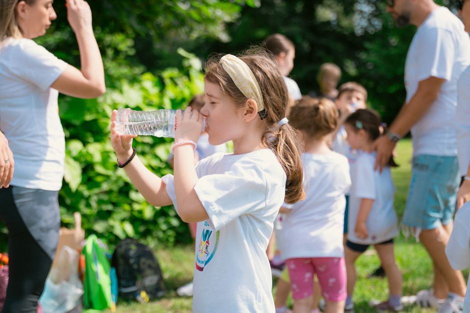 Young girl drinking water during sunny outdoor activity with friends in a park setting.